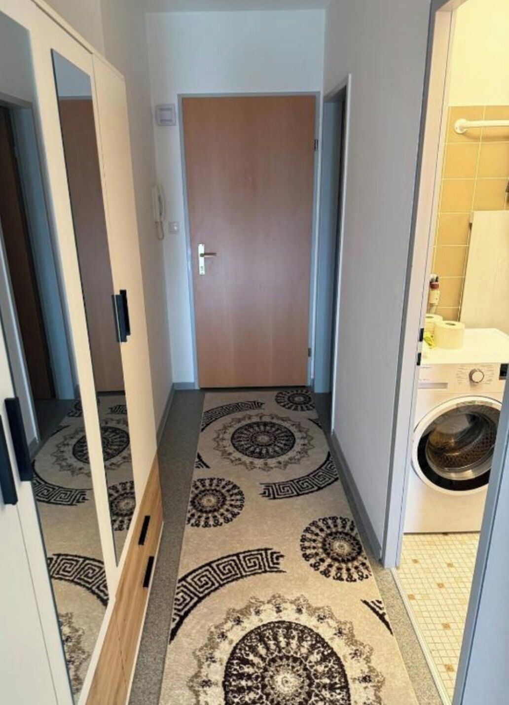 A narrow hallway with a patterned carpet leading to a door, a washing machine in the adjacent room, and a tiled floor.
