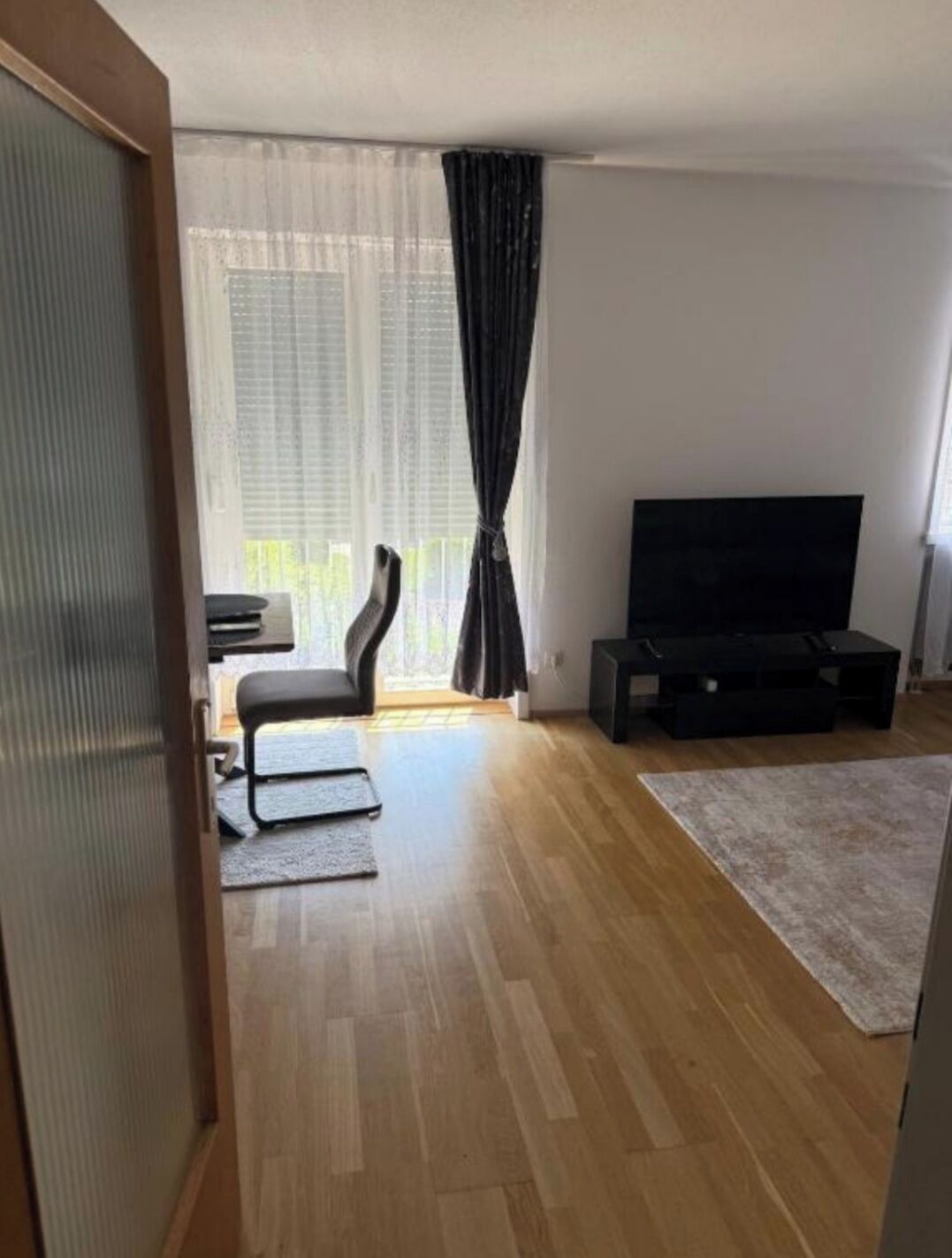 Ein Wohnzimmer mit Flachbildfernseher, einer Couch, einem Couchtisch und einem großen Fenster mit Vorhängen.