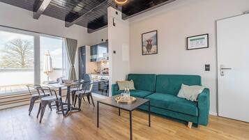 Ein modernes Wohnzimmer mit einem grünen Sofa, einem Esstisch und Stühlen. Im Hintergrund ist eine Küche mit einem großen Fenster zu sehen.