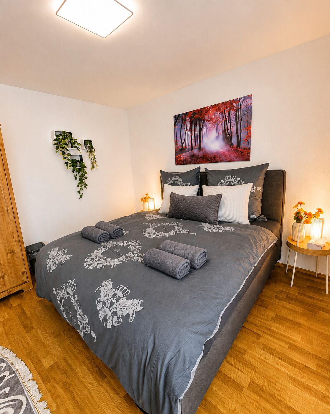 A neatly made bed with a grey bedspread and rolled towels, a painting of a forest scene on the wall, and a small table with a vase of flowers.