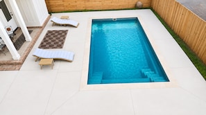 A rectangular swimming pool with a built-in hot tub, surrounded by a white tiled deck, a wooden fence, and a small garden area with a bench and a patterned rug.