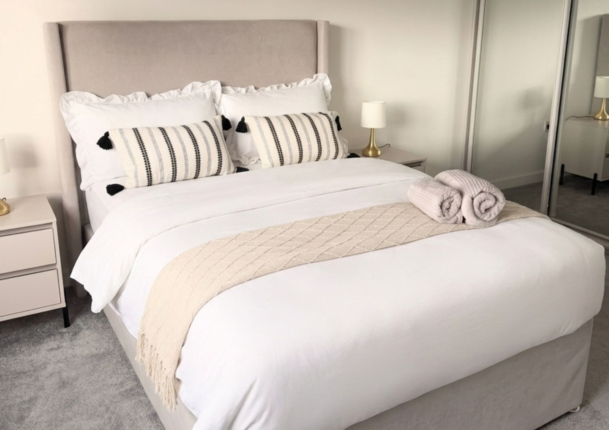 A neatly made bed with white bedding and a beige throw blanket, a bedside lamp, and a nightstand with a lamp.