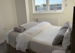 En enkeltseng med hvide sengetøj og et mønstret tæppe, et vindue med udsigt til bygninger og en radiator på væggen.