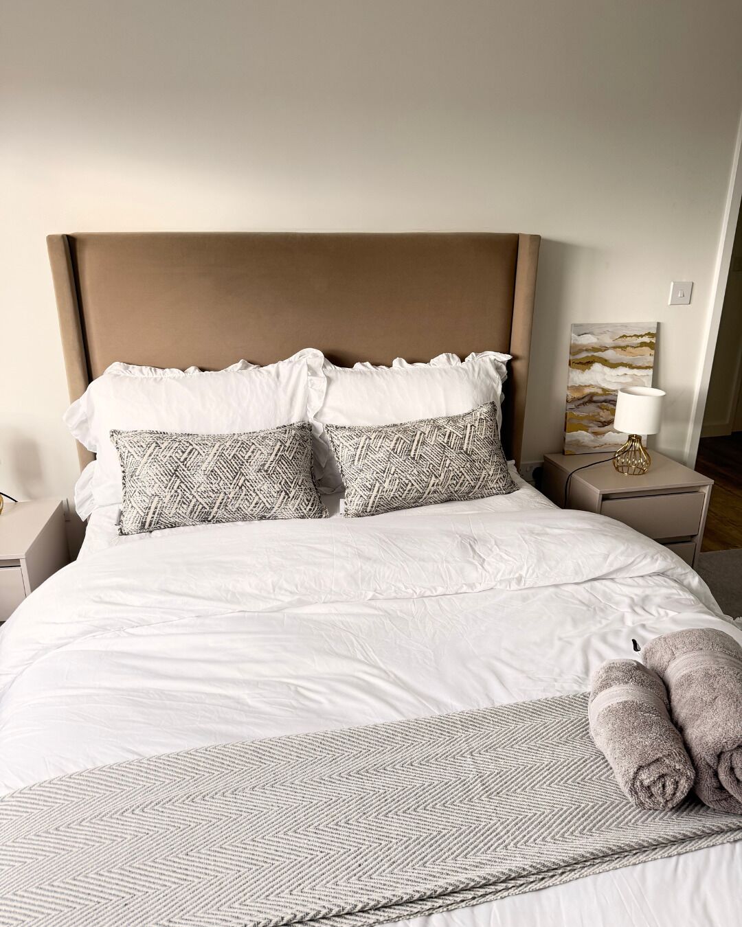 A neatly made bed with white bedding and patterned pillows, a bedside lamp, and a wall art piece.