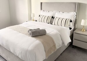 Un lit bien fait, avec une tête de lit grise, une literie blanche et un plaid beige. Deux tables de chevet, chacune surmontée d’une lampe, se trouvent de part et d’autre du lit.