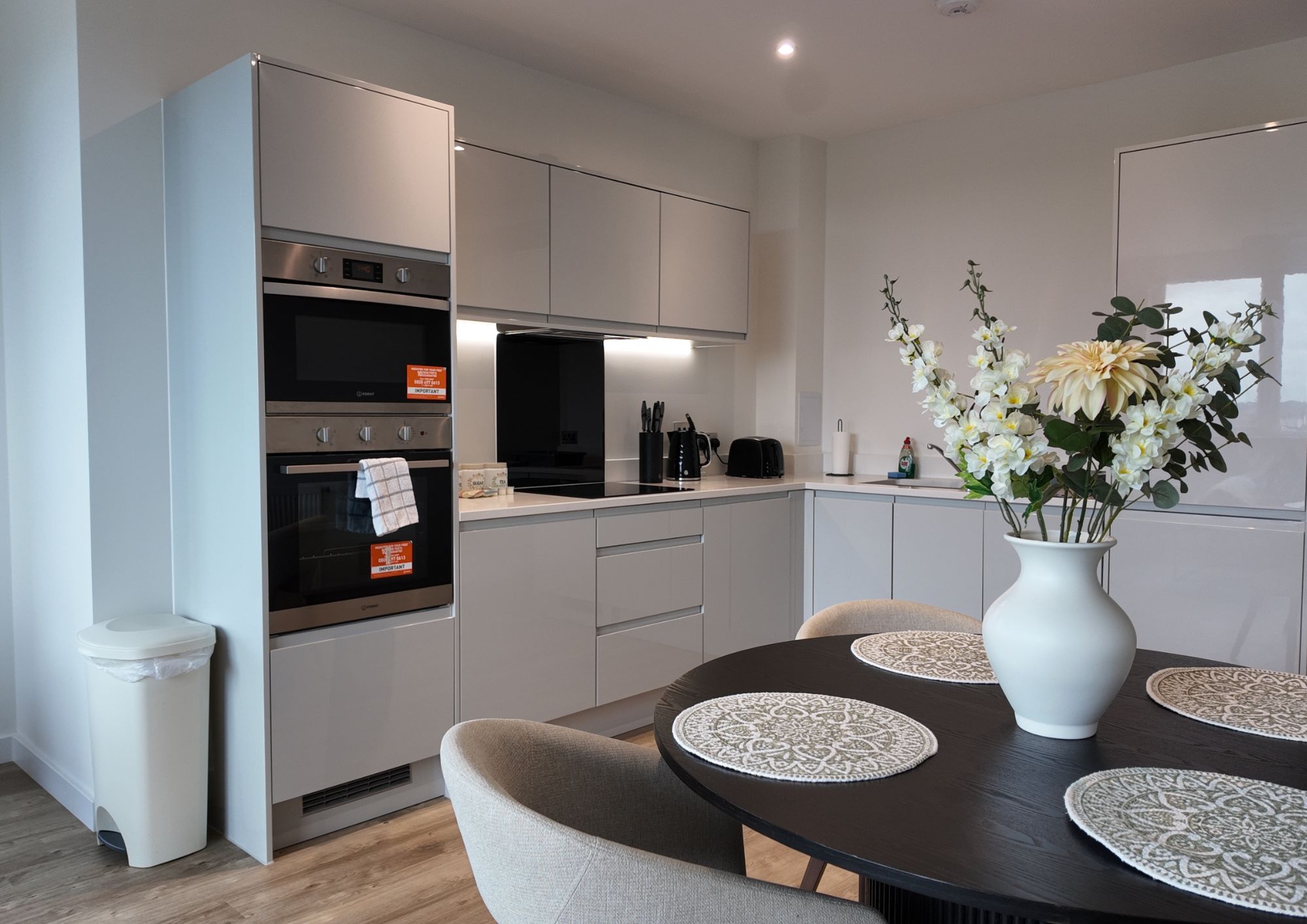 A modern kitchen with built-in appliances, a dining table with placemats, and a vase with flowers.