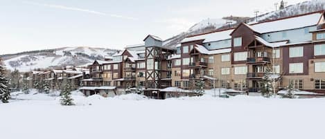 A snow-covered resort with multi-story buildings, ski slopes in the background, and a mountainous landscape.