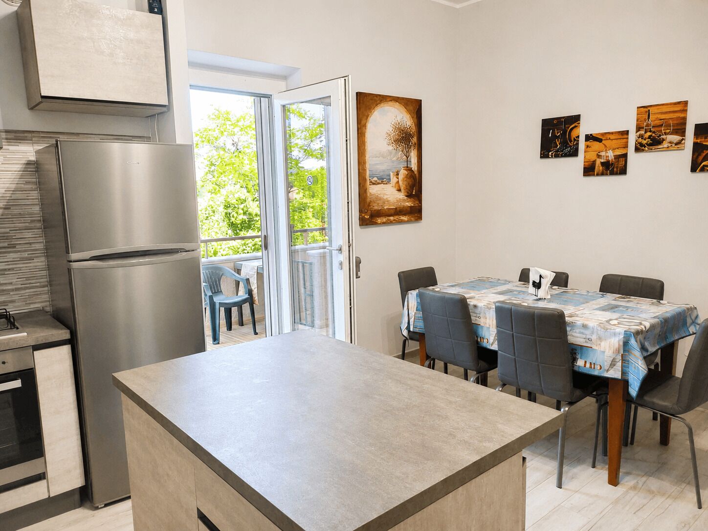 A modern kitchen with a built-in refrigerator, a dining area with a table and chairs, and a view through the open door to an outdoor area with greenery.