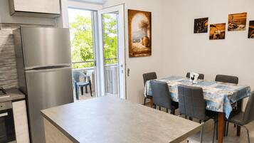 A modern kitchen with a built-in refrigerator, a dining area with a table and chairs, and a view through the open door to an outdoor area with greenery.