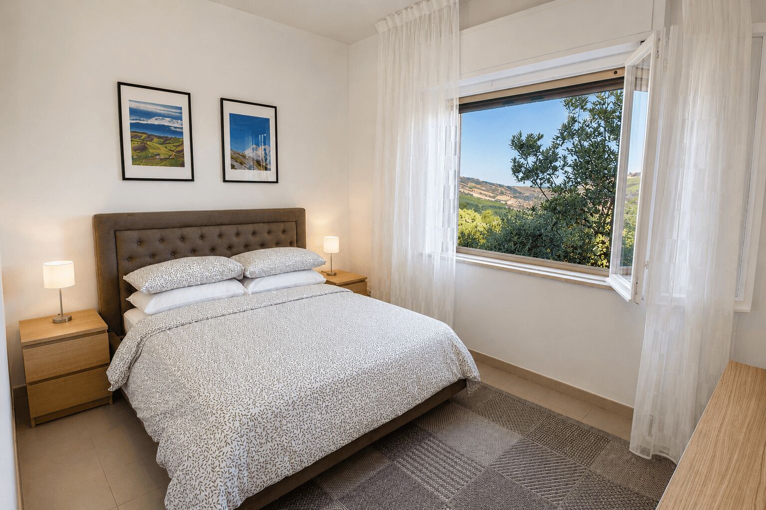 A bedroom with a bed, two bedside tables, a window with a view, and two framed pictures on the wall.