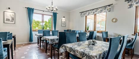 A dining area with multiple tables and chairs, a chandelier, and windows with curtains.