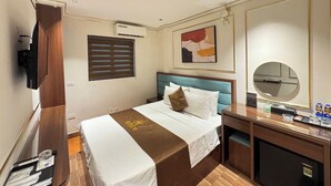 Une chambre d’hôtel équipée d’un lit, d’un bureau, d’un miroir, d’une télévision et d’une fenêtre avec des stores.