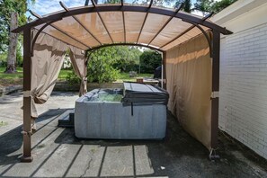 Ein überdachter Außenbereich mit Whirlpool unter einer Markise, eine Ziegelwand und ein Garten im Hintergrund.
