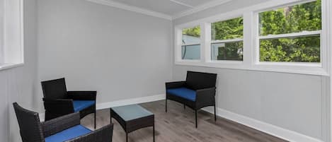 Une pièce avec deux fauteuils en osier et une petite table, des murs blancs et de grandes fenêtres donnant sur un paysage verdoyant.