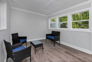 Ein Zimmer mit zwei Korbstühlen, einem kleinen Tisch, weißen Wänden und großen Fenstern mit Blick auf Grünflächen.