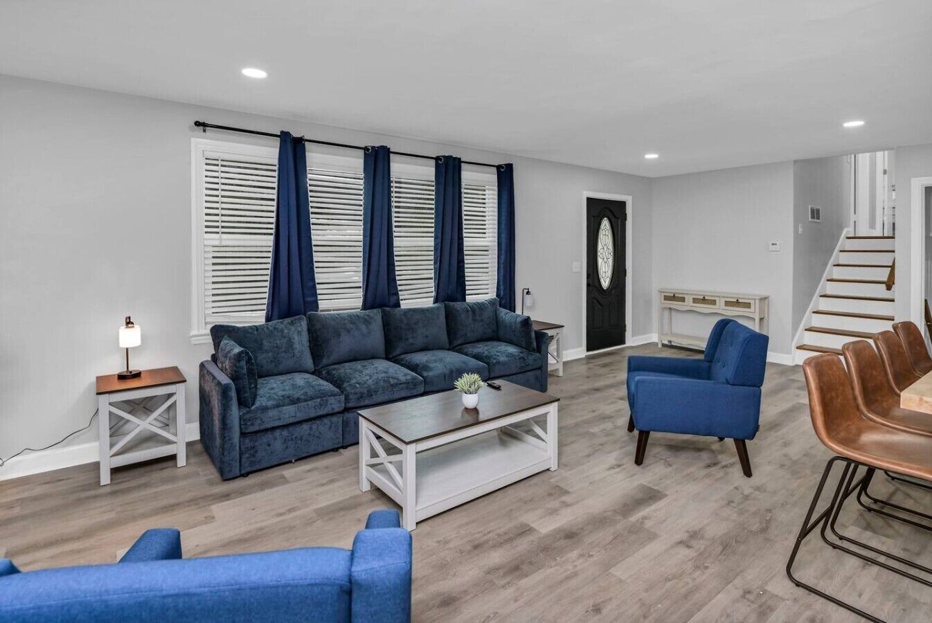 Un salon moderne avec un canapé d’angle, une table basse, un fauteuil bleu, une table à manger avec des chaises, et un escalier en arrière-plan.