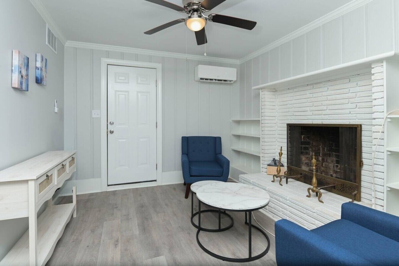 A room with a white door, a blue chair, a round table, a fireplace, and a ceiling fan.