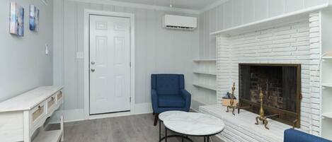 A room with a white door, a blue chair, a round table, a fireplace, and a ceiling fan.