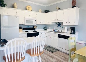A compact kitchen with white cabinets, a microwave, a dishwasher, and a dining area with two white chairs. - 124 Seashell Cove Poinciana Place (Miramar Beach)