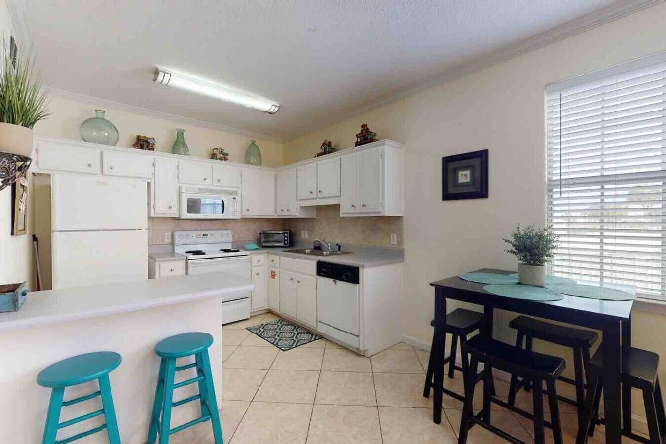Cocina compacta con gabinetes blancos, un microondas, estufa y pileta. Hay dos taburetes y una mesa con sillas.