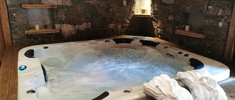 Un bain à remous avec un mur en pierre en arrière-plan, des étagères en bois et des serviettes posées sur le sol.