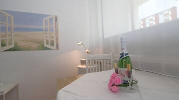 A room with a bed, a table with a bottle and glasses, a chair, a painting of a beach, and a window with blinds.