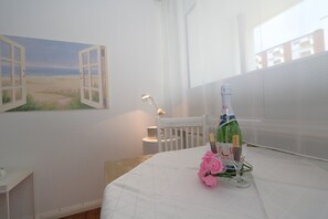 Ein Zimmer mit einem Bett, einem Tisch mit Flasche und Gläsern, einem Stuhl, einem Bild einer Strandansicht und einem Fenster mit Jalousien.