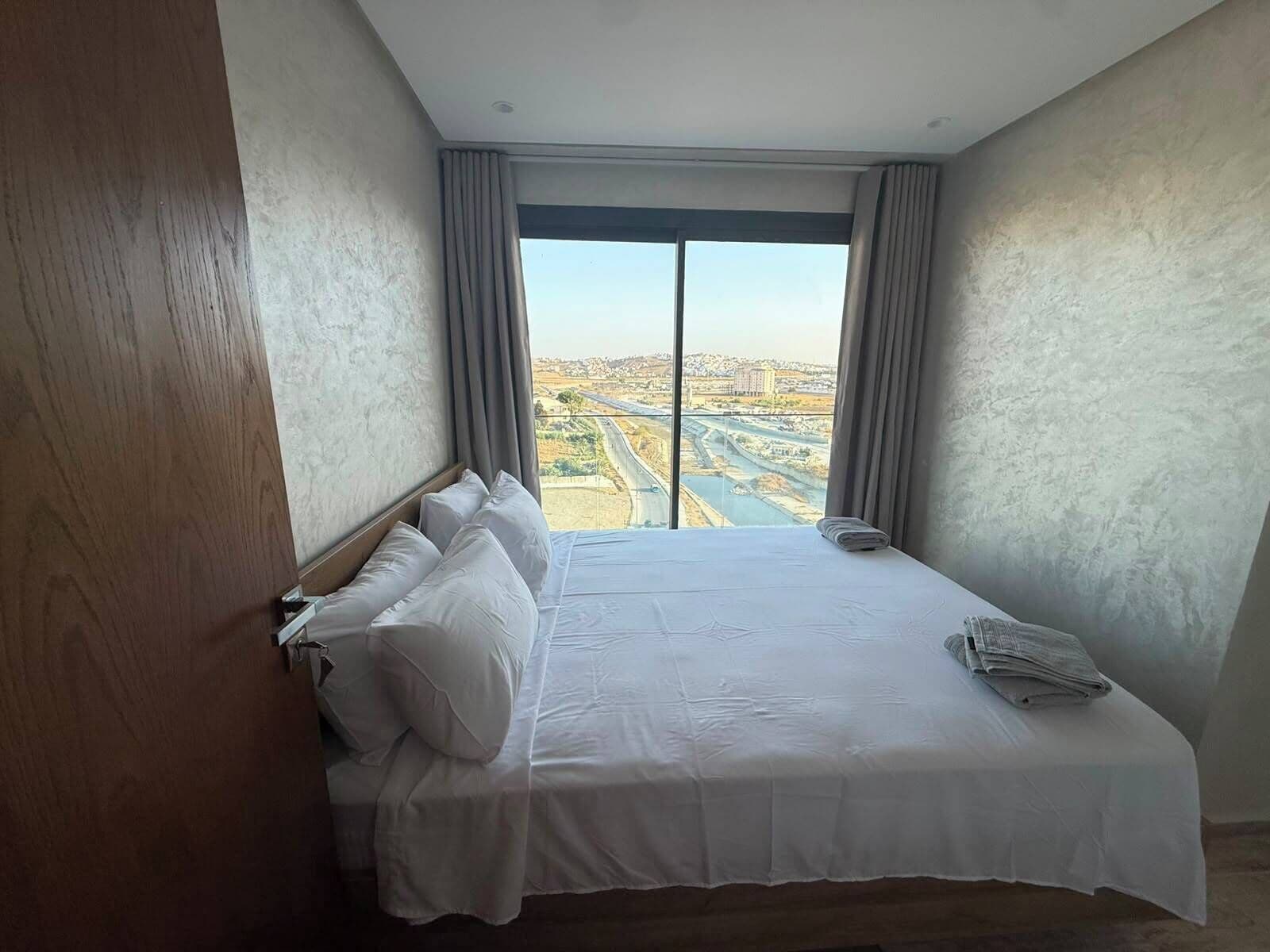 A hotel room with a large bed, a view of a cityscape, and a wooden door.