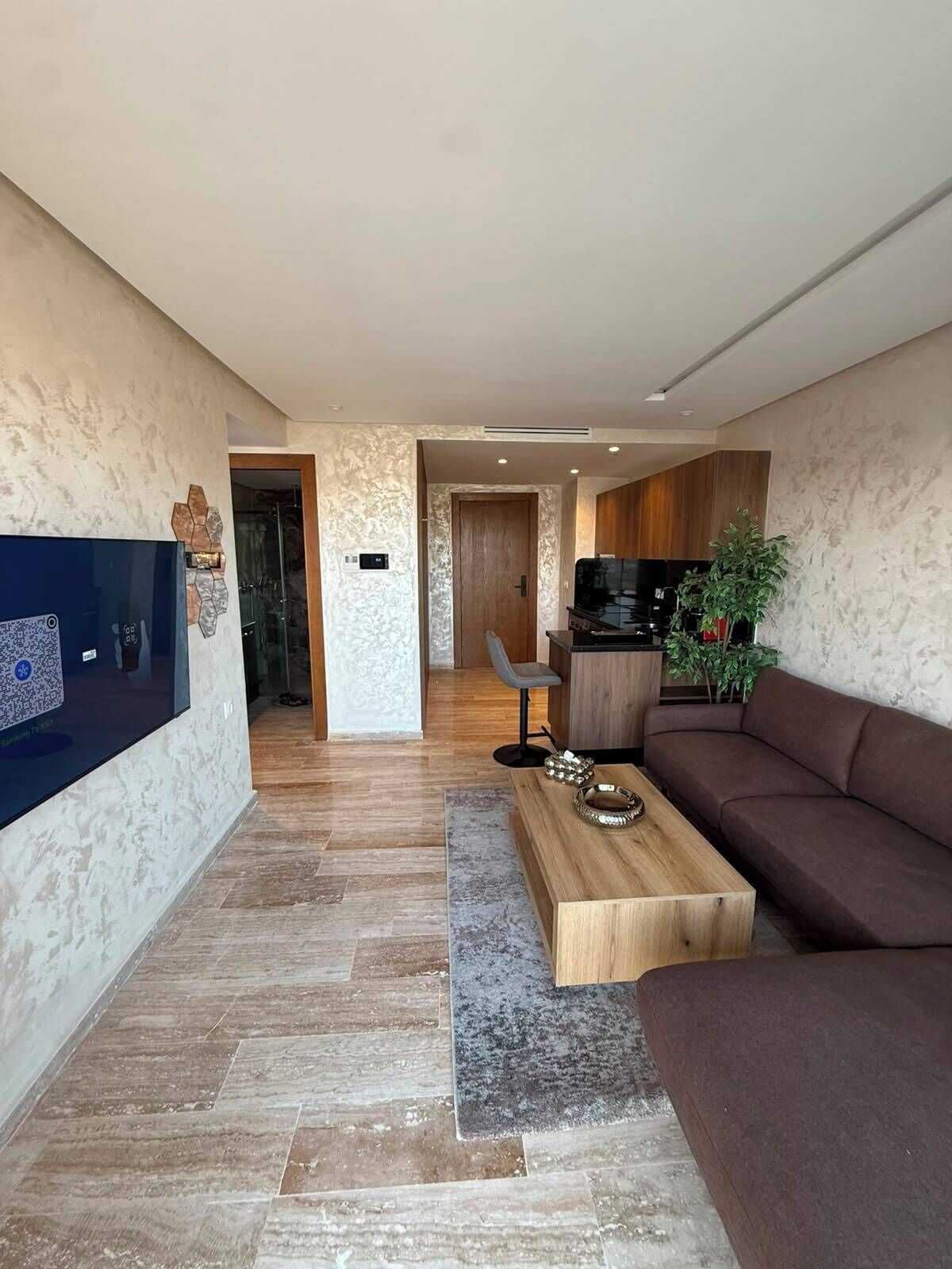 Un salon moderne avec un téléviseur à écran plat, un canapé marron, une table basse en bois et un coin cuisine à l’arrière-plan.