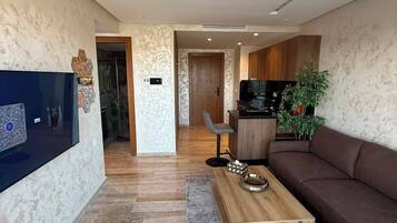A modern living room with a flat-screen TV, a brown sofa, a wooden coffee table, and a kitchen area in the background.