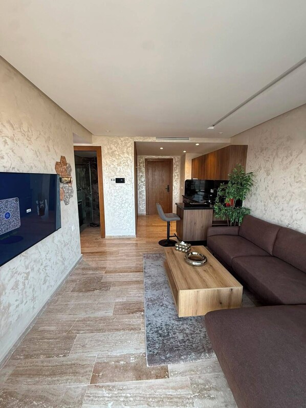 A modern living room with a flat-screen TV, a brown sofa, a wooden coffee table, and a kitchen area in the background.