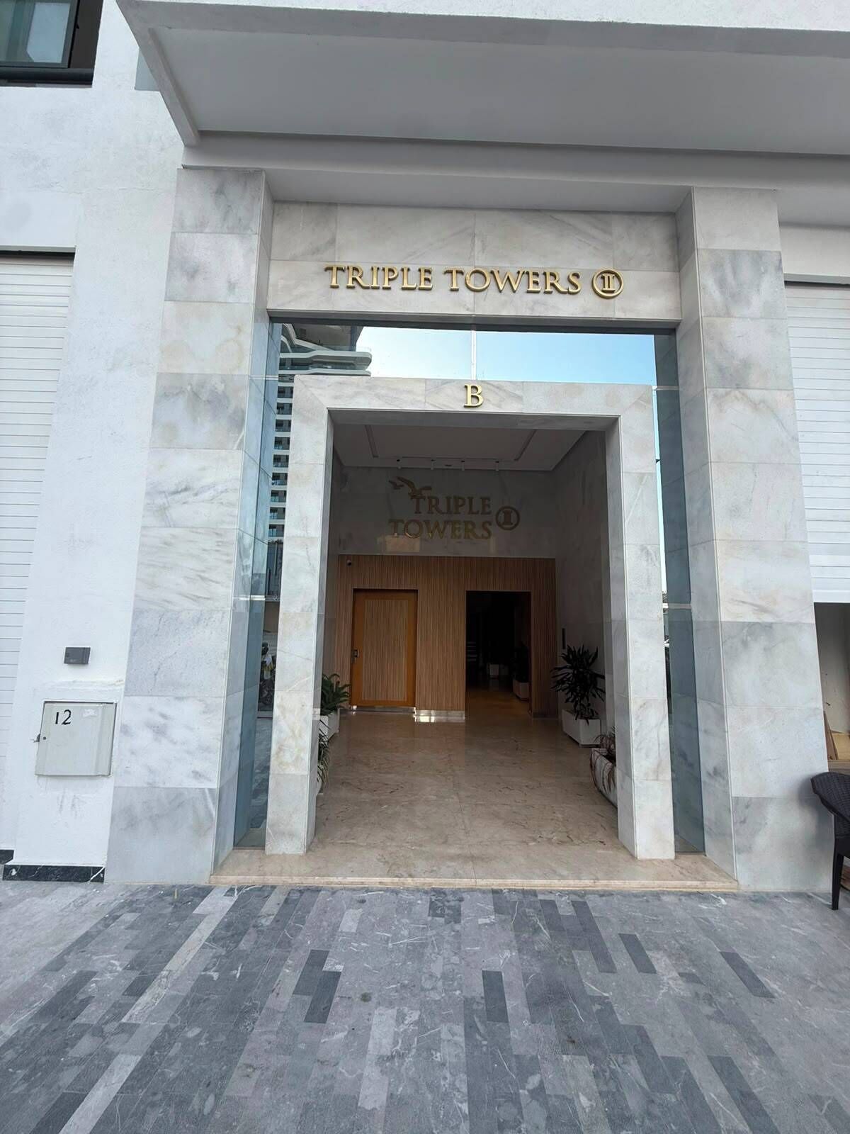 The entrance to Triple Towers with a marble facade, a wooden door, and a clear sky above.