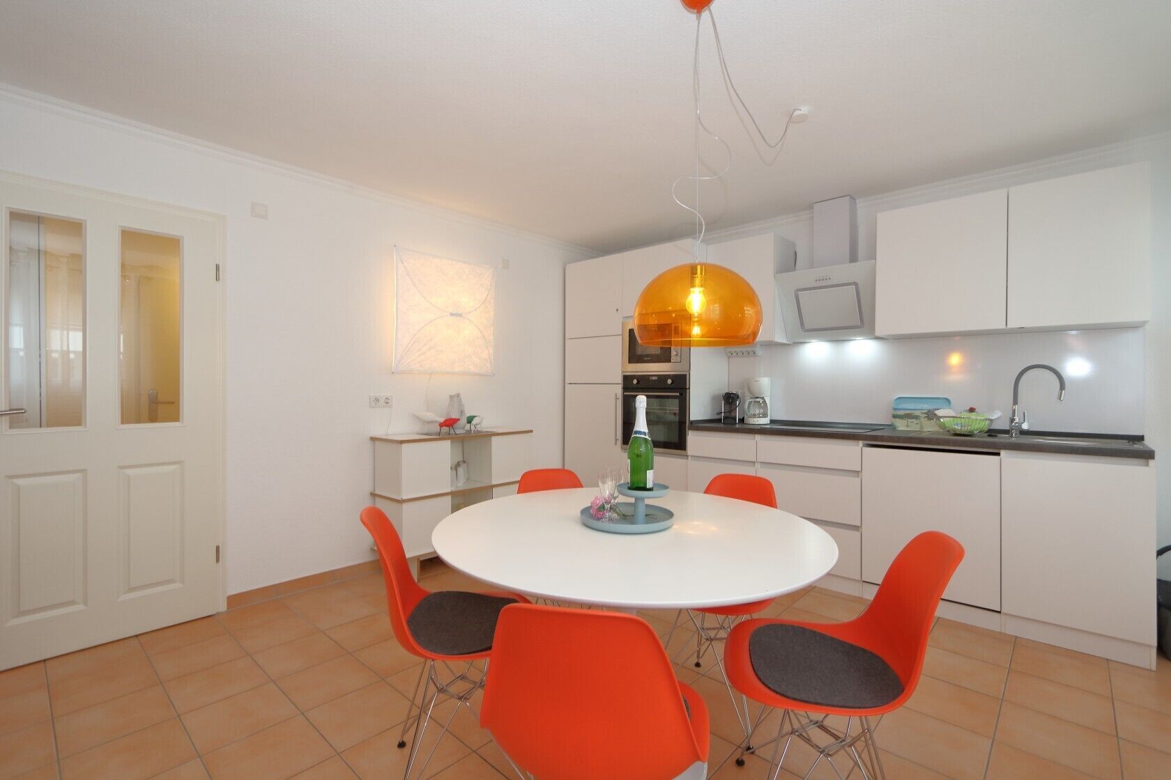 A modern kitchen with white cabinets, a round dining table with orange chairs, and a pendant light.