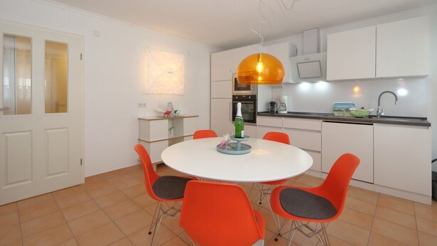 Een moderne keuken met witte kastjes, een ronde eettafel met oranje stoelen en een hanglamp.