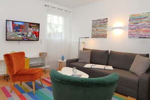 Ein modernes Wohnzimmer mit einer Couch, Sesseln, einem Couchtisch, einem Fernseher und einem Fenster mit Vorhängen.