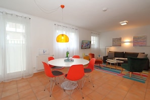 A modern living room with a round dining table, orange chairs, a green armchair, a sofa, and a colorful rug. - App. Silvie 4 <br>Just a few meters from the beach access, the “Schwalbe” vacation rental is located on the mezzanine level of the Winora building in Westerland. <br>The one-bedroom apartment is approximately 38 m2 in size (Westerland)
