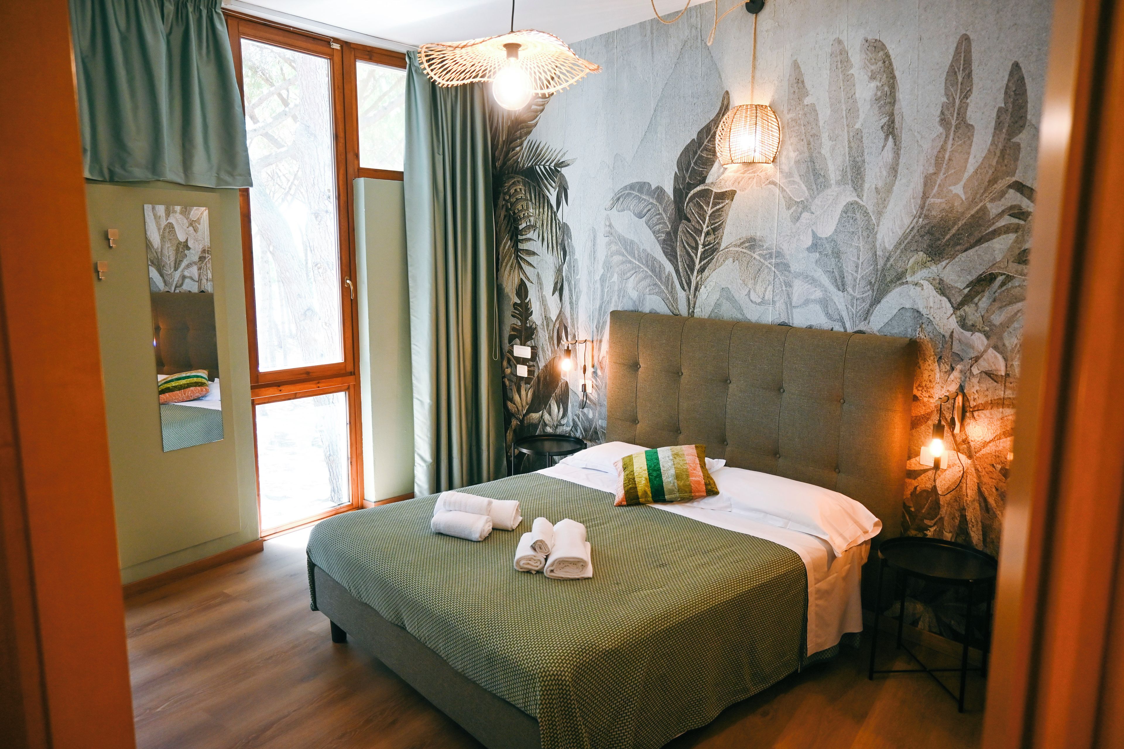 Une chambre d’hôtel avec un grand lit, une fenêtre avec des rideaux, un mur orné d’un motif de feuilles et un luminaire suspendu.