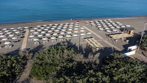 Vista aérea de uma praia com fileiras de espreguiçadeiras e guarda-sóis, um deck de madeira e um pequeno edifício.