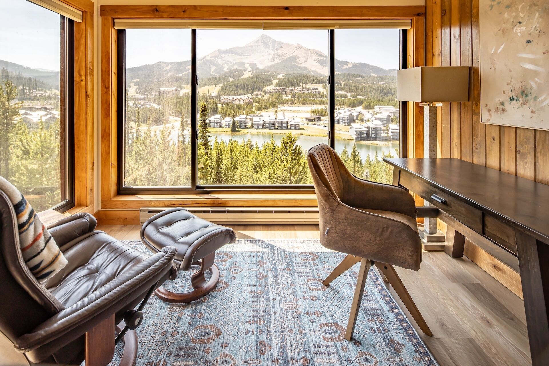 A room with a large window, a leather sofa, a wooden desk, and a patterned rug.