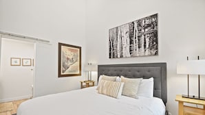 Un dormitorio con una cama grande, un cabecero tapizado en gris, una mesita de noche con una lámpara y un cuadro en la pared.