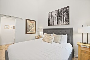 Ein Schlafzimmer mit einem großen Bett, einem grauen, gepolsterten Kopfteil, einem Nachttisch mit Lampe und einem gerahmten Bild an der Wand.