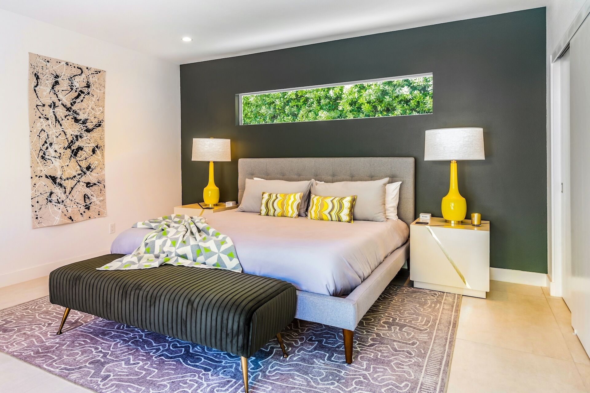 Une chambre moderne avec un lit, deux tables de chevet avec des lampes, un grand tableau abstrait et une fenêtre donnant sur un paysage verdoyant.