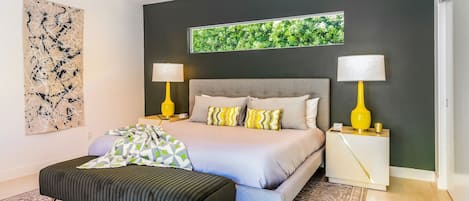 Une chambre moderne avec un lit, deux tables de chevet avec des lampes, un grand tableau abstrait et une fenêtre donnant sur un paysage verdoyant.