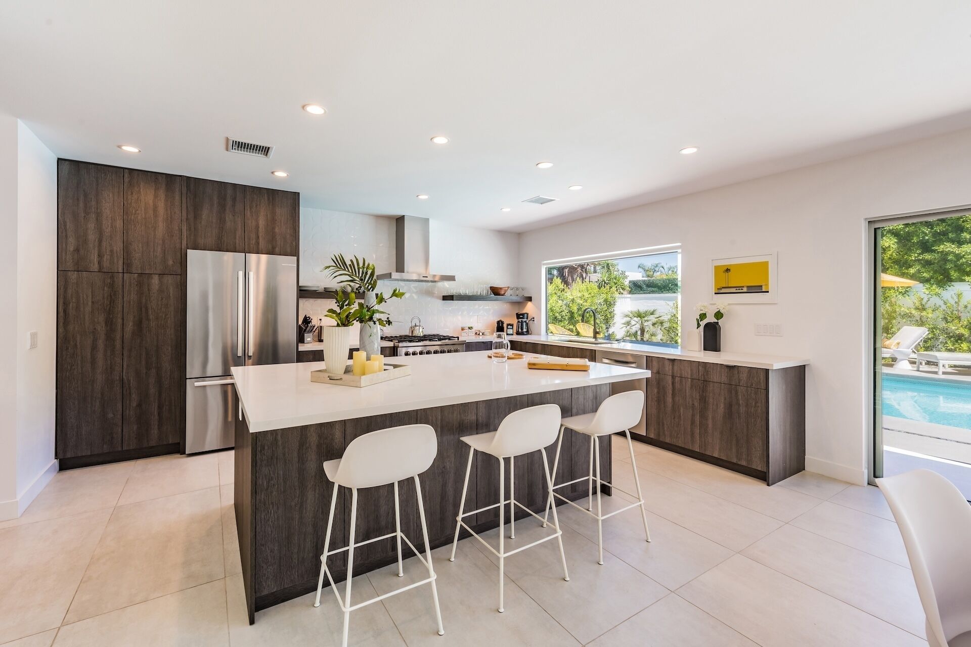 A modern kitchen with a central island, bar stools, a stainless steel refrigerator, and a view of a swimming pool.