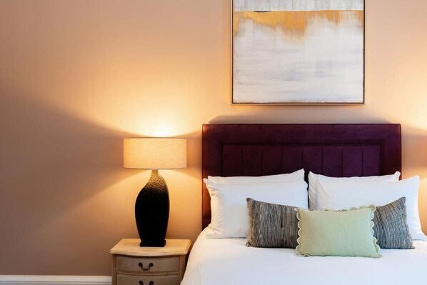 Ein ordentlich bezogenes Bett mit Kopfteil, einer Nachttischlampe und einem an der Wand hängenden abstrakten Gemälde.