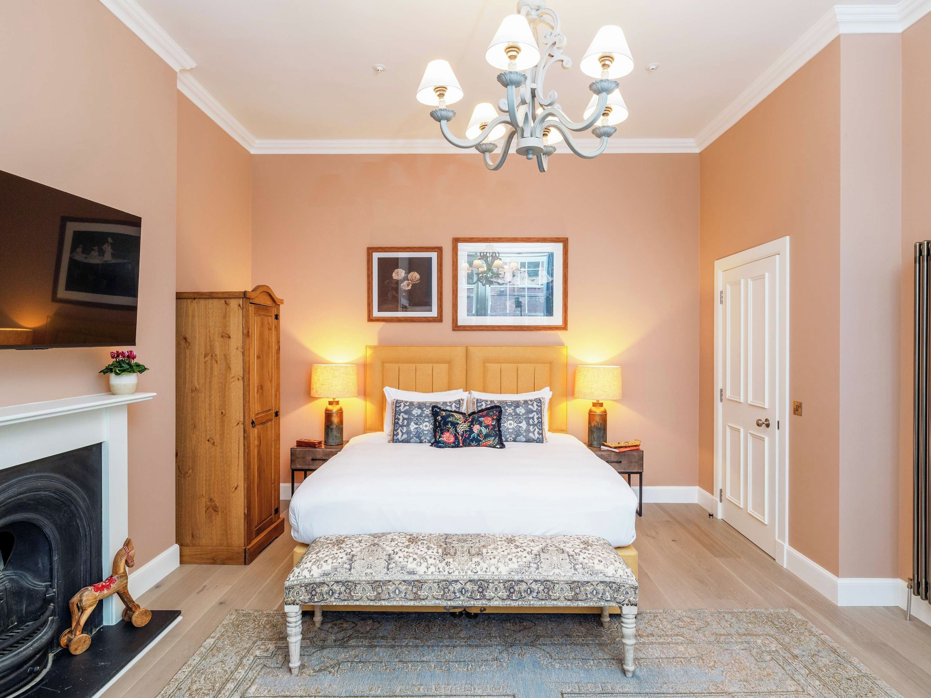 A bedroom with a bed, bedside lamps, a chandelier, a fireplace, and a door.