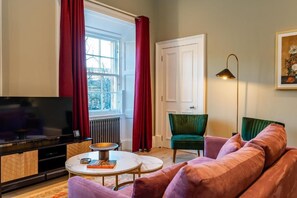 Ein Wohnzimmer mit Fernseher, rundem Couchtisch, Sofa und Sessel. Ein Fenster mit Vorhängen, eine Heizung und eine Tür sind ebenfalls vorhanden.