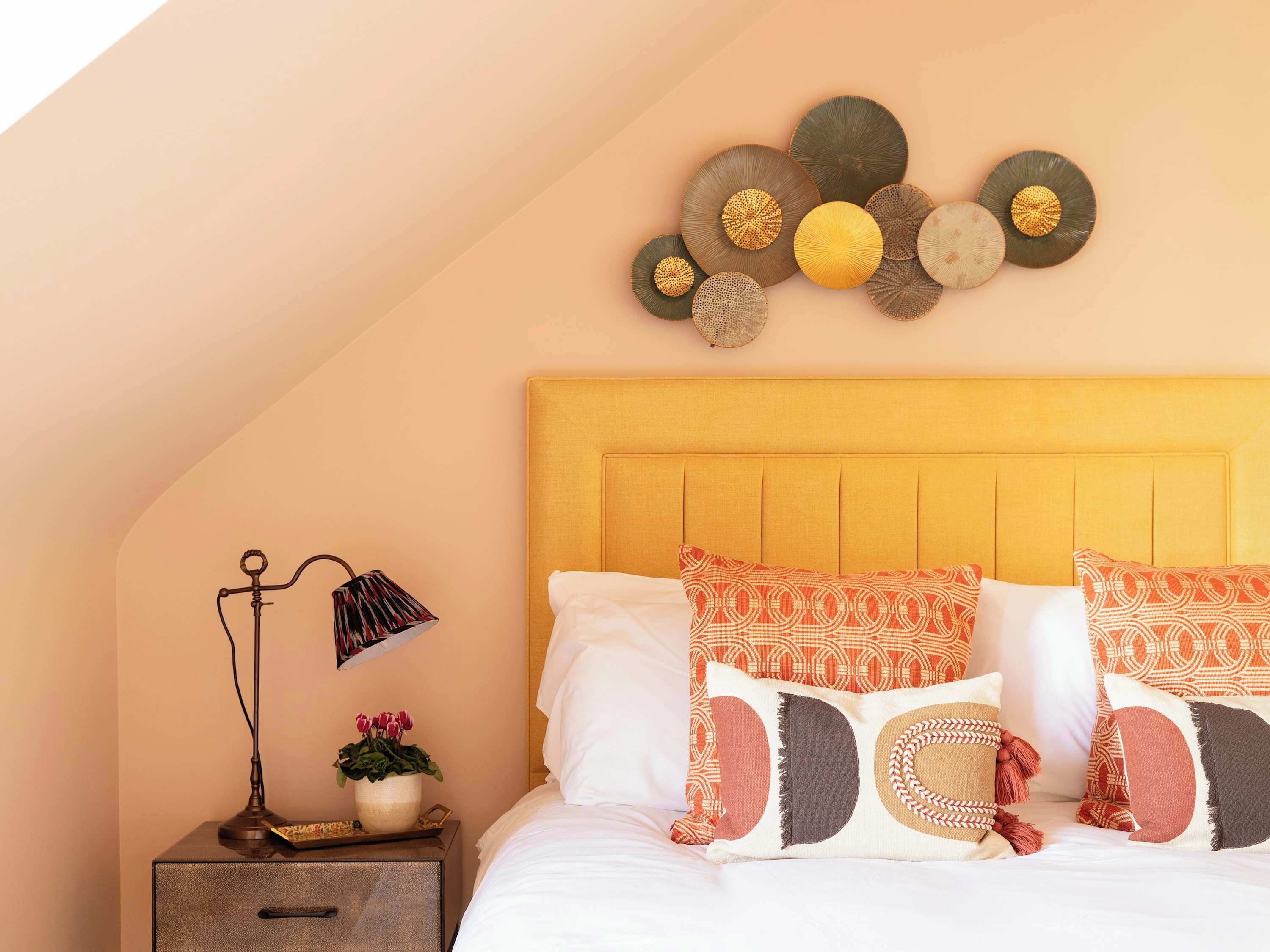 Quarto com cama, almofadas, mesa de cabeceira, luminária e decoração na parede.