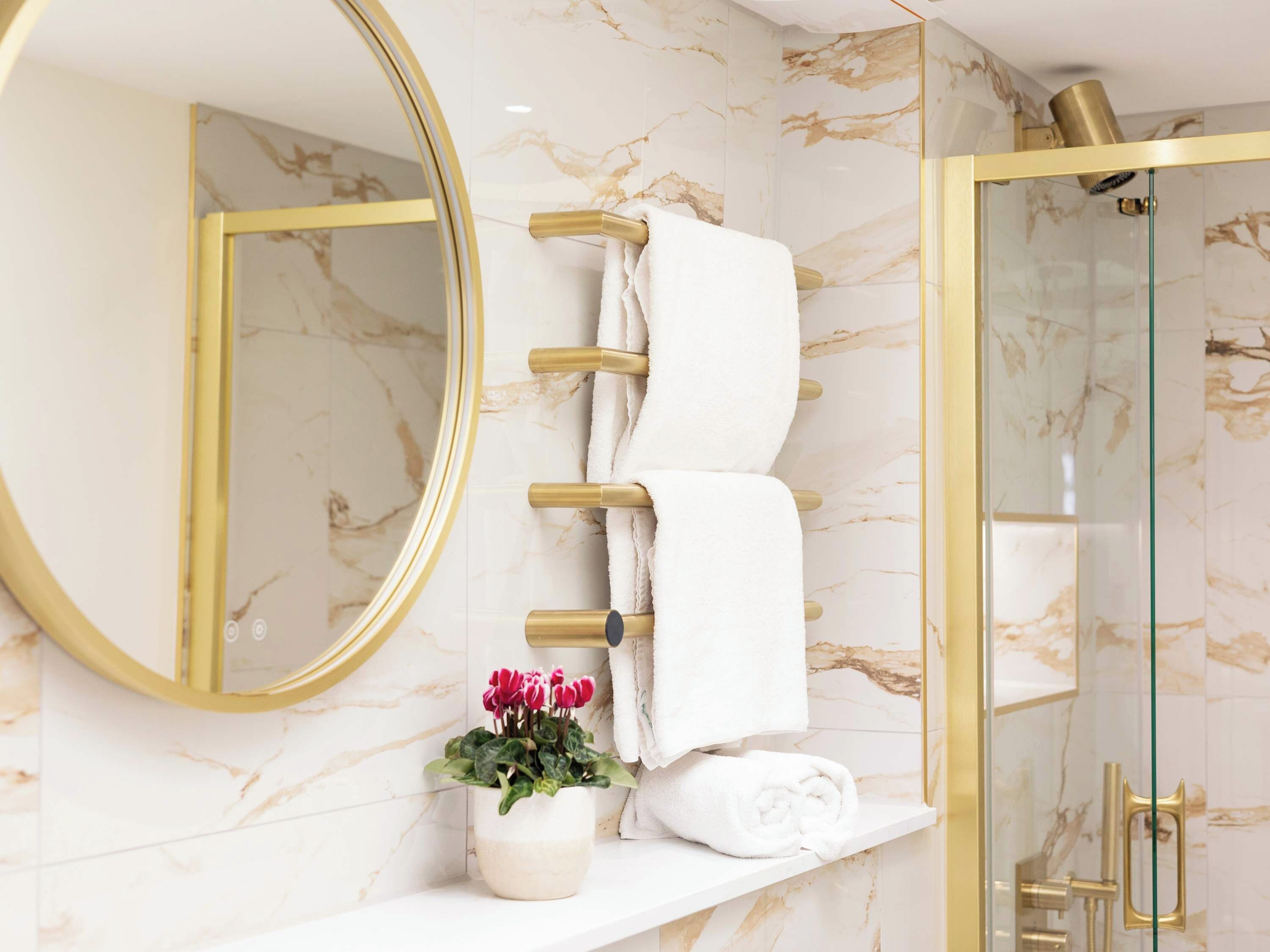 Un baño con pared de mármol, espejo con marco dorado, toallero, planta en maceta y cabina de ducha de vidrio.