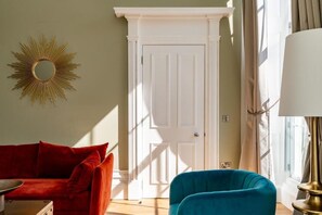 Ein Zimmer mit einem roten Sofa, einem blauen Sessel, einem goldfarbenen Spiegel und einer weißen Tür.